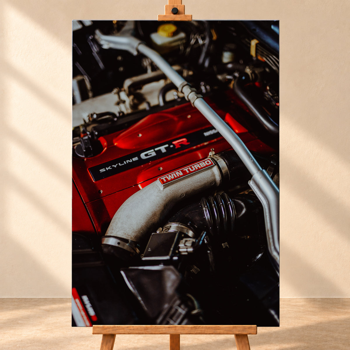 Nahaufnahme eines Motorraums mit roter Abdeckung und sichtbarem SKYLINE GT-R-Schriftzug, hellem Rohr mit TWIN TURBO-Kennzeichnung, schwarzer Verrohrung und einer silbernen, diagonal verlaufenden Strebe; dunkler Hintergrund mit warmen Lichtreflexen; Visualisierung als Leinwand-Wandbild, frontal auf einer Holzstaffelei.