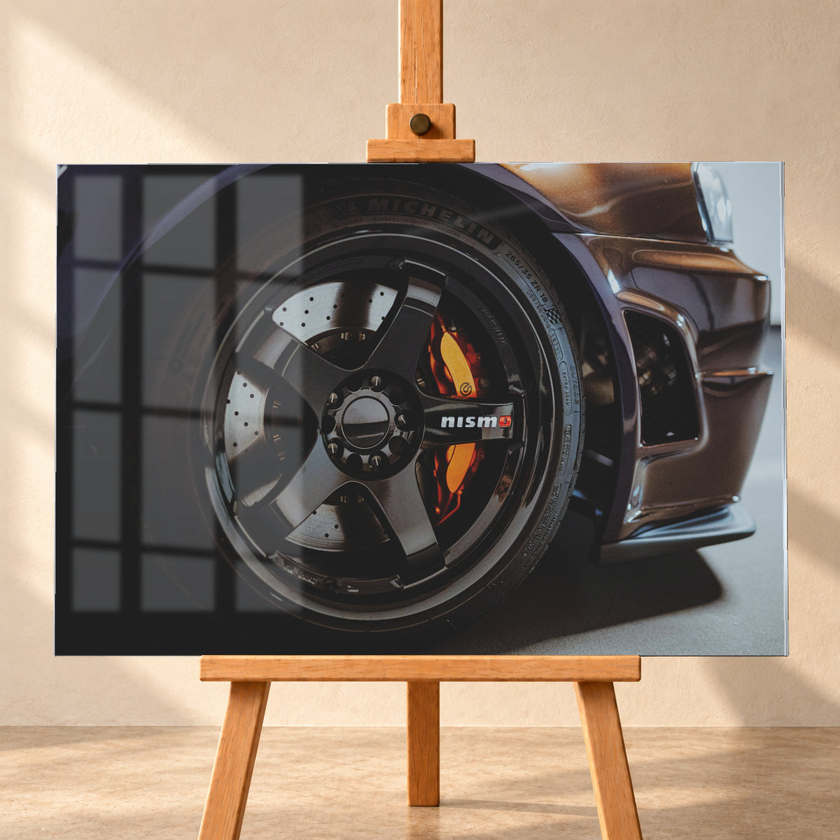 Nahaufnahme der Frontpartie eines dunkelviolett-bronze schimmernden Nissan Skyline R34 mit schwarzem Rad, sichtbarem NISMO-Schriftzug, gelochter Bremsscheibe und orange-rotem Bremssattel bei kontrastreichem Licht; Visualisierung als Acrylglas-Wandbild, frontal auf einer Holzstaffelei.