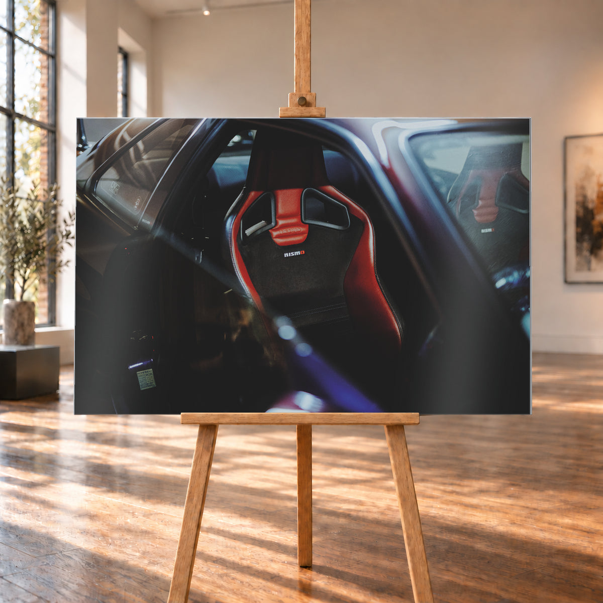 Seitlicher Nahblick in den dunklen Innenraum eines Nissan Skyline R34. Ein rot-schwarzer Sportsitz mit Öffnungen in der Rückenlehne und Nismo-Schriftzug steht im Zentrum; ein zweiter Sitz ist gegenüber sichtbar. Blau-violette Reflexe, Glasflächen und tiefe Schatten rahmen die Szene; Visualisierung als Alu gebürstet-Wandbild, auf einer Holzstaffelei in einem hellen Galerieraum.