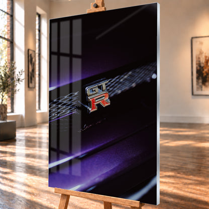 Nahaufnahme eines rot-silbernen GT-R-Emblems an einem dunklen Frontgrill, beleuchtet von violettem Licht und umgeben von tiefen Schatten; Visualisierung als Acrylglas-Wandbild, schräg von der Seite auf einer Holzstaffelei.