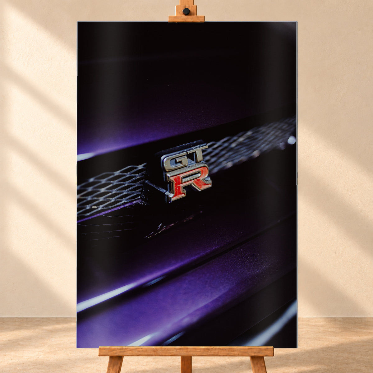 Nahaufnahme eines rot-silbernen GT-R-Emblems an einem dunklen Frontgrill, beleuchtet von violettem Licht und umgeben von tiefen Schatten; Visualisierung als Alu gebürstet-Wandbild, frontal auf einer Holzstaffelei.