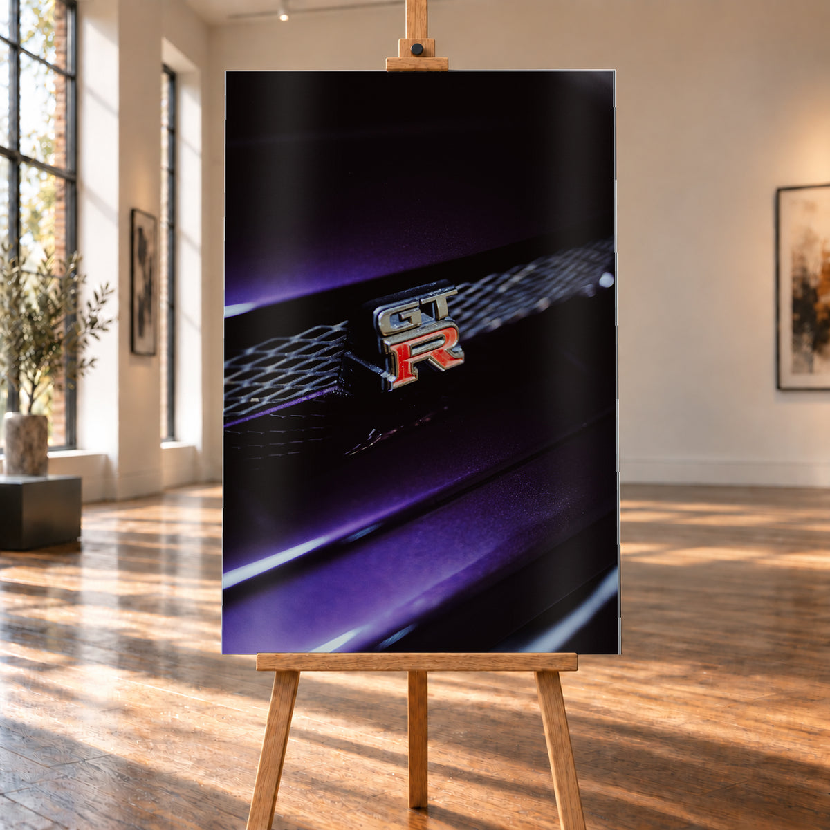 Nahaufnahme eines rot-silbernen GT-R-Emblems an einem dunklen Frontgrill, beleuchtet von violettem Licht und umgeben von tiefen Schatten; Visualisierung als Alu gebürstet-Wandbild, auf einer Holzstaffelei in einem hellen Galerieraum.
