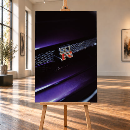 Nahaufnahme eines rot-silbernen GT-R-Emblems an einem dunklen Frontgrill, beleuchtet von violettem Licht und umgeben von tiefen Schatten; Visualisierung als Leinwand-Wandbild, auf einer Holzstaffelei in einem hellen Galerieraum.