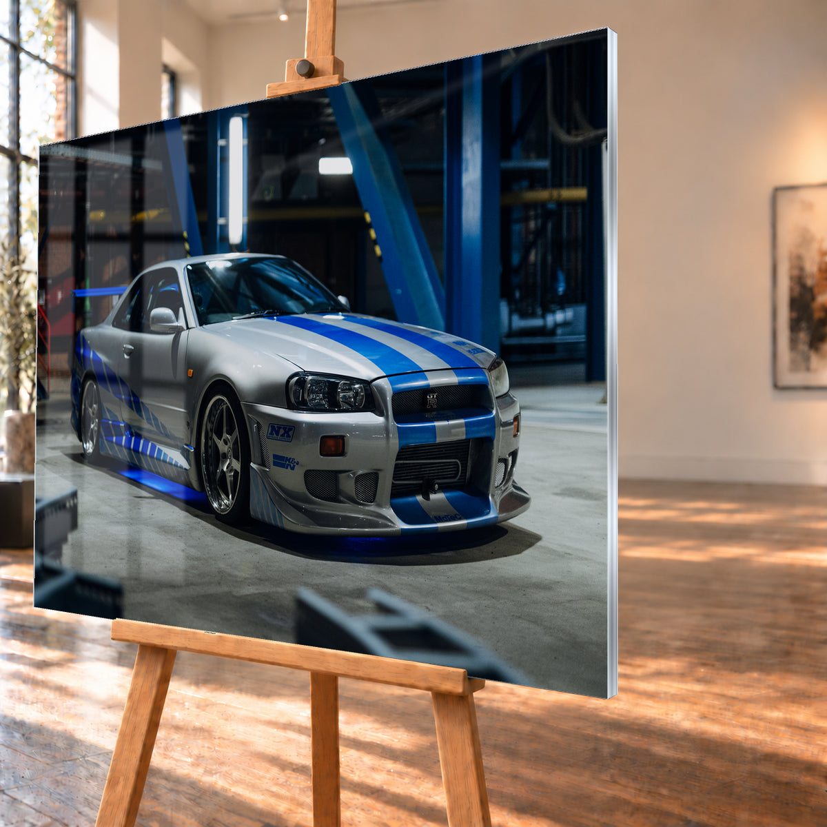 Silberner Nissan Skyline GT-R R34 mit blauen Streifen und Seitengrafik in tiefer Front-Dreiviertelansicht. Das Fahrzeug steht auf Beton in einer dunklen Industriehalle mit blauen Stahlträgern, hellen Leuchten und blauem Licht unter der Karosserie. Frontschürze und großer Heckflügel sind sichtbar; Visualisierung als Acrylglas-Wandbild, schräg von der Seite auf einer Holzstaffelei.