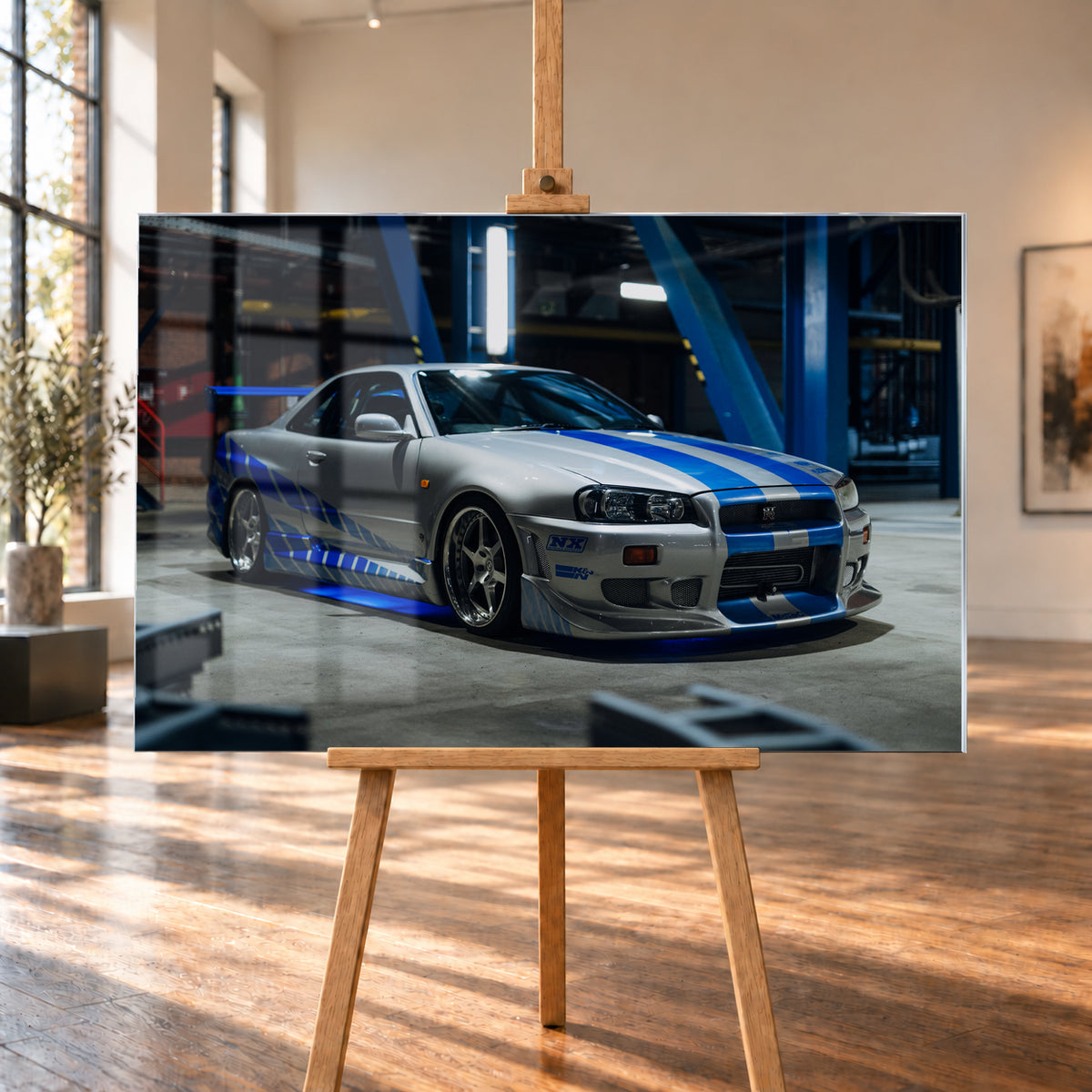 Silberner Nissan Skyline GT-R R34 mit blauen Streifen und Seitengrafik in tiefer Front-Dreiviertelansicht. Das Fahrzeug steht auf Beton in einer dunklen Industriehalle mit blauen Stahlträgern, hellen Leuchten und blauem Licht unter der Karosserie. Frontschürze und großer Heckflügel sind sichtbar; Visualisierung als Acrylglas-Wandbild, auf einer Holzstaffelei in einem hellen Galerieraum.