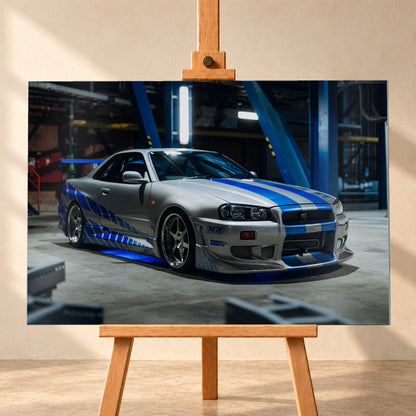 Silberner Nissan Skyline GT-R R34 mit blauen Streifen und Seitengrafik in tiefer Front-Dreiviertelansicht. Das Fahrzeug steht auf Beton in einer dunklen Industriehalle mit blauen Stahlträgern, hellen Leuchten und blauem Licht unter der Karosserie. Frontschürze und großer Heckflügel sind sichtbar; Visualisierung als Alu gebürstet-Wandbild, frontal auf einer Holzstaffelei.