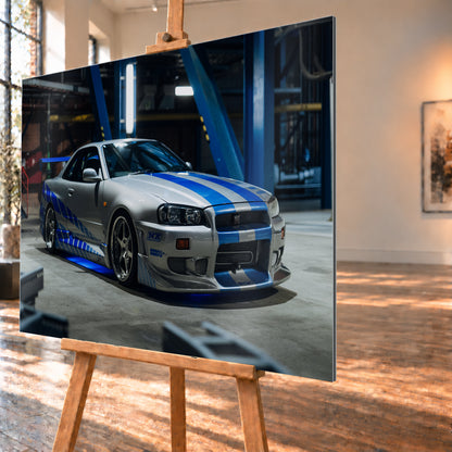 Silberner Nissan Skyline GT-R R34 mit blauen Streifen und Seitengrafik in tiefer Front-Dreiviertelansicht. Das Fahrzeug steht auf Beton in einer dunklen Industriehalle mit blauen Stahlträgern, hellen Leuchten und blauem Licht unter der Karosserie. Frontschürze und großer Heckflügel sind sichtbar; Visualisierung als Alu gebürstet-Wandbild, schräg von der Seite auf einer Holzstaffelei.