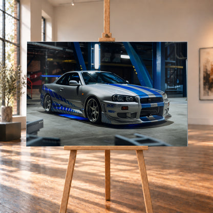 Silberner Nissan Skyline GT-R R34 mit blauen Streifen und Seitengrafik in tiefer Front-Dreiviertelansicht. Das Fahrzeug steht auf Beton in einer dunklen Industriehalle mit blauen Stahlträgern, hellen Leuchten und blauem Licht unter der Karosserie. Frontschürze und großer Heckflügel sind sichtbar; Visualisierung als Alu gebürstet-Wandbild, auf einer Holzstaffelei in einem hellen Galerieraum.