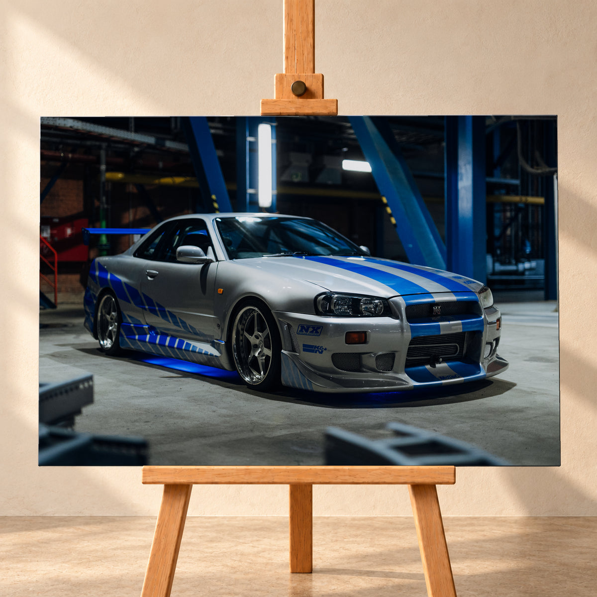 Silberner Nissan Skyline GT-R R34 mit blauen Streifen und Seitengrafik in tiefer Front-Dreiviertelansicht. Das Fahrzeug steht auf Beton in einer dunklen Industriehalle mit blauen Stahlträgern, hellen Leuchten und blauem Licht unter der Karosserie. Frontschürze und großer Heckflügel sind sichtbar; Visualisierung als Leinwand-Wandbild, frontal auf einer Holzstaffelei.