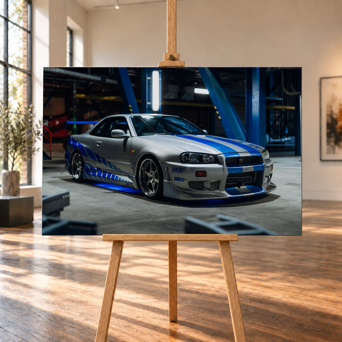 Silberner Nissan Skyline GT-R R34 mit blauen Streifen und Seitengrafik in tiefer Front-Dreiviertelansicht. Das Fahrzeug steht auf Beton in einer dunklen Industriehalle mit blauen Stahlträgern, hellen Leuchten und blauem Licht unter der Karosserie. Frontschürze und großer Heckflügel sind sichtbar; Visualisierung als Leinwand-Wandbild, auf einer Holzstaffelei in einem hellen Galerieraum.