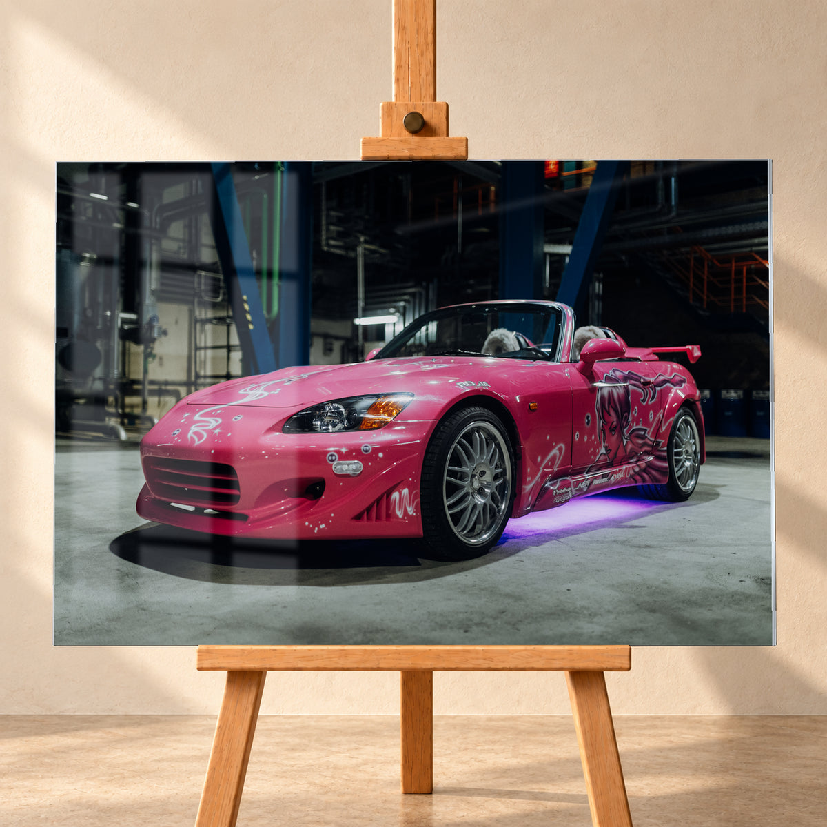 Pinker Honda S2000 als offener Roadster in Front-Dreiviertelansicht auf einem Betonboden in einer dunklen Industriehalle; sichtbar sind weiße Dekorgrafiken, eine große Frauenfigur an der Seite, silberne Mehrspeichenräder, ein Heckflügel und violett-blaues Licht unter dem Fahrzeug; Visualisierung als Acrylglas-Wandbild, frontal auf einer Holzstaffelei.