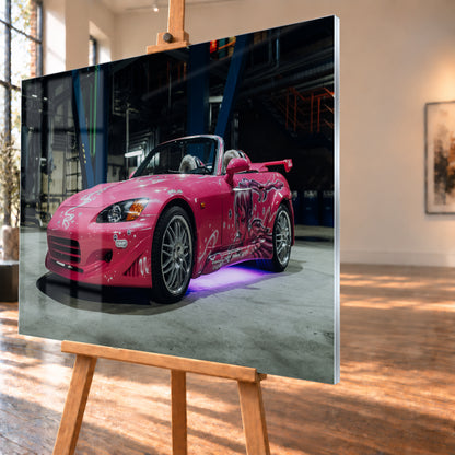 Pinker Honda S2000 als offener Roadster in Front-Dreiviertelansicht auf einem Betonboden in einer dunklen Industriehalle; sichtbar sind weiße Dekorgrafiken, eine große Frauenfigur an der Seite, silberne Mehrspeichenräder, ein Heckflügel und violett-blaues Licht unter dem Fahrzeug; Visualisierung als Acrylglas-Wandbild, schräg von der Seite auf einer Holzstaffelei.