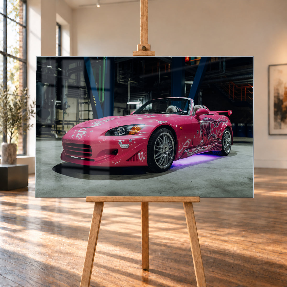 Pinker Honda S2000 als offener Roadster in Front-Dreiviertelansicht auf einem Betonboden in einer dunklen Industriehalle; sichtbar sind weiße Dekorgrafiken, eine große Frauenfigur an der Seite, silberne Mehrspeichenräder, ein Heckflügel und violett-blaues Licht unter dem Fahrzeug; Visualisierung als Acrylglas-Wandbild, auf einer Holzstaffelei in einem hellen Galerieraum.