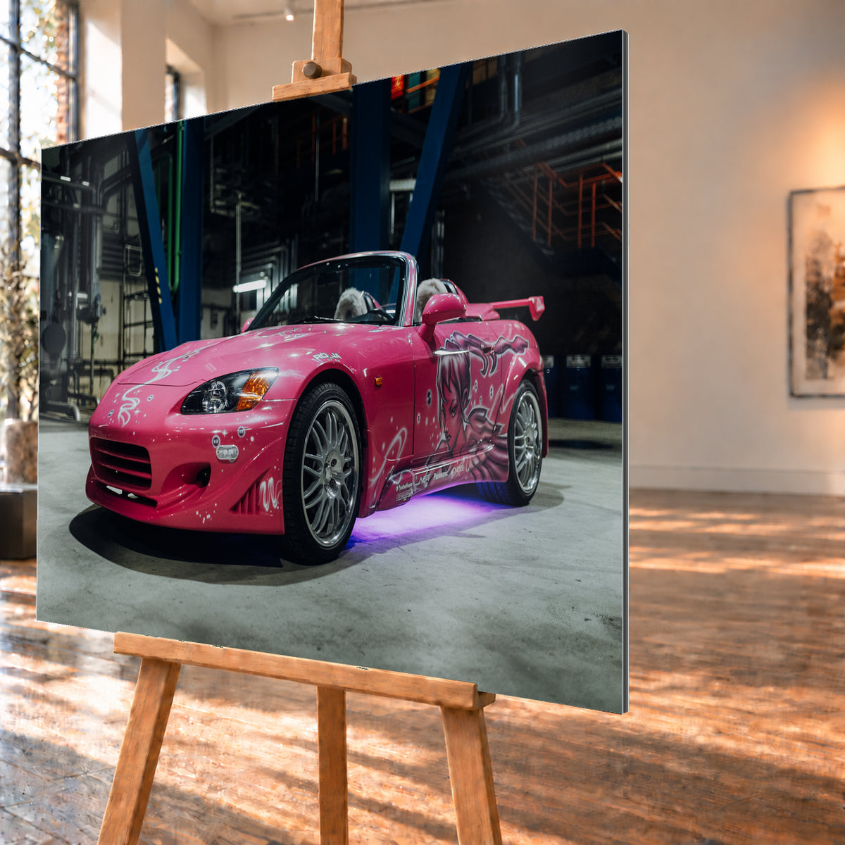 Pinker Honda S2000 als offener Roadster in Front-Dreiviertelansicht auf einem Betonboden in einer dunklen Industriehalle; sichtbar sind weiße Dekorgrafiken, eine große Frauenfigur an der Seite, silberne Mehrspeichenräder, ein Heckflügel und violett-blaues Licht unter dem Fahrzeug; Visualisierung als Alu gebürstet-Wandbild, schräg von der Seite auf einer Holzstaffelei.