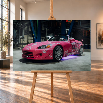 Pinker Honda S2000 als offener Roadster in Front-Dreiviertelansicht auf einem Betonboden in einer dunklen Industriehalle; sichtbar sind weiße Dekorgrafiken, eine große Frauenfigur an der Seite, silberne Mehrspeichenräder, ein Heckflügel und violett-blaues Licht unter dem Fahrzeug; Visualisierung als Alu gebürstet-Wandbild, auf einer Holzstaffelei in einem hellen Galerieraum.