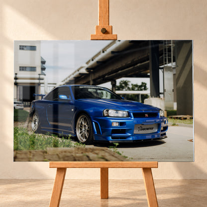 Blauer Nissan Skyline R34 GT-R in tiefer Front-Dreiviertelansicht auf Asphalt unter einer erhöhten Betonstruktur; sichtbar sind silberne Mehrspeichenräder, Heckflügel, GT-R-Emblem und Gras am Bildrand; Visualisierung als Acrylglas-Wandbild, frontal auf einer Holzstaffelei.