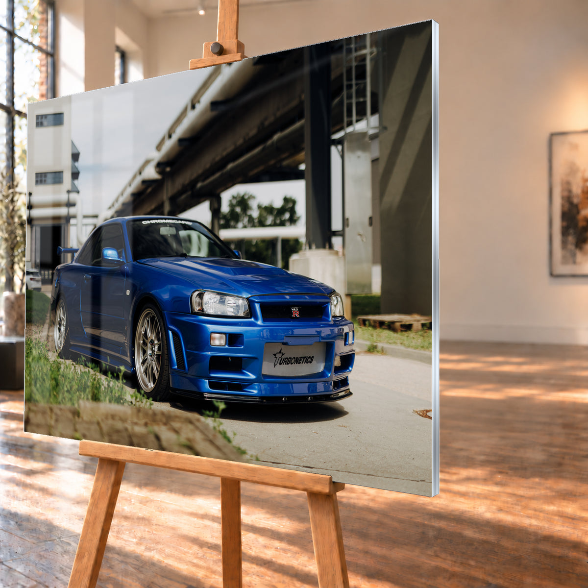 Blauer Nissan Skyline R34 GT-R in tiefer Front-Dreiviertelansicht auf Asphalt unter einer erhöhten Betonstruktur; sichtbar sind silberne Mehrspeichenräder, Heckflügel, GT-R-Emblem und Gras am Bildrand; Visualisierung als Acrylglas-Wandbild, schräg von der Seite auf einer Holzstaffelei.