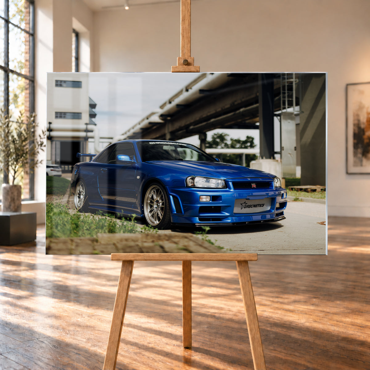 Blauer Nissan Skyline R34 GT-R in tiefer Front-Dreiviertelansicht auf Asphalt unter einer erhöhten Betonstruktur; sichtbar sind silberne Mehrspeichenräder, Heckflügel, GT-R-Emblem und Gras am Bildrand; Visualisierung als Acrylglas-Wandbild, auf einer Holzstaffelei in einem hellen Galerieraum.