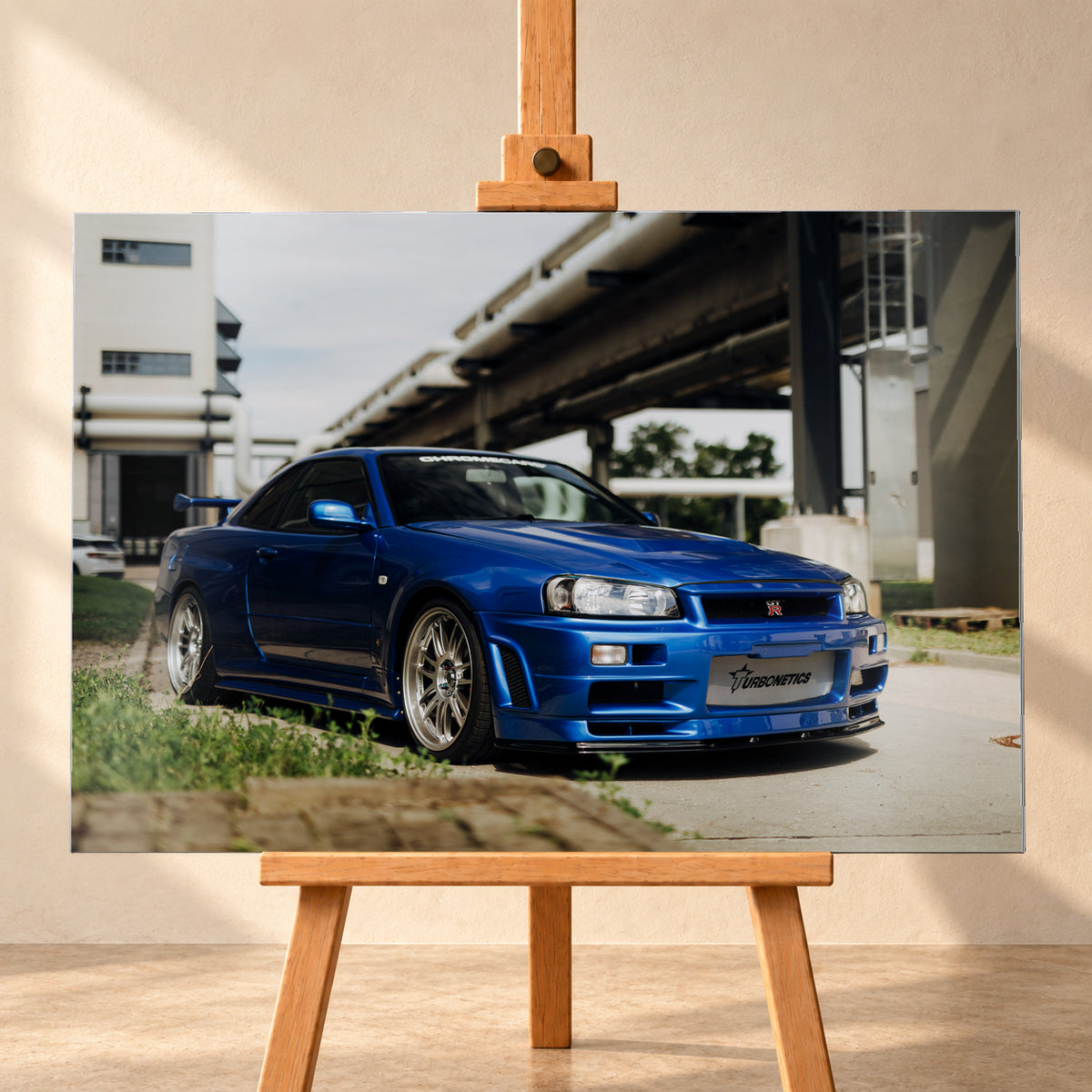 Blauer Nissan Skyline R34 GT-R in tiefer Front-Dreiviertelansicht auf Asphalt unter einer erhöhten Betonstruktur; sichtbar sind silberne Mehrspeichenräder, Heckflügel, GT-R-Emblem und Gras am Bildrand; Visualisierung als Alu gebürstet-Wandbild, frontal auf einer Holzstaffelei.