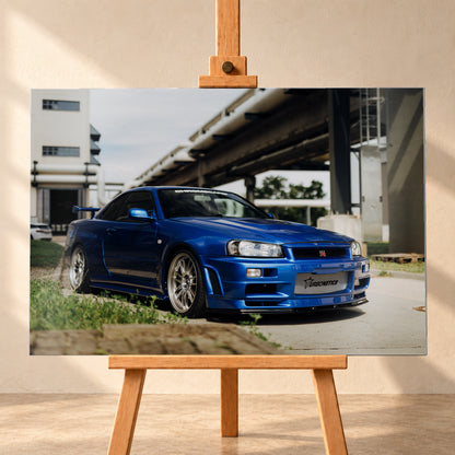 Blauer Nissan Skyline R34 GT-R in tiefer Front-Dreiviertelansicht auf Asphalt unter einer erhöhten Betonstruktur; sichtbar sind silberne Mehrspeichenräder, Heckflügel, GT-R-Emblem und Gras am Bildrand; Visualisierung als Alu gebürstet-Wandbild, frontal auf einer Holzstaffelei.