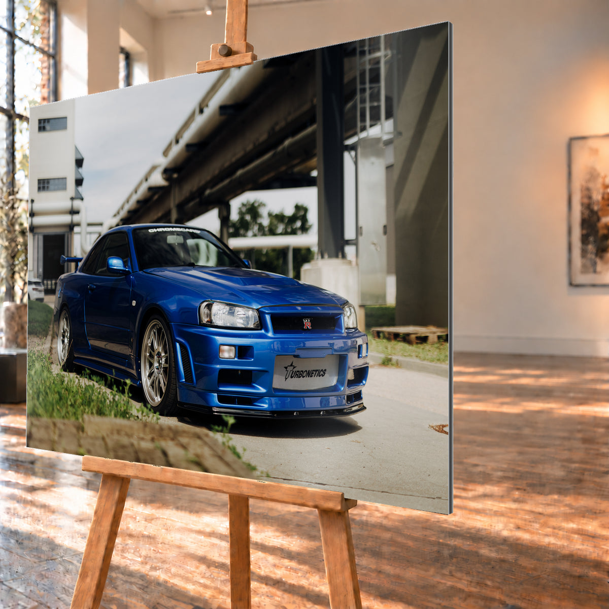 Blauer Nissan Skyline R34 GT-R in tiefer Front-Dreiviertelansicht auf Asphalt unter einer erhöhten Betonstruktur; sichtbar sind silberne Mehrspeichenräder, Heckflügel, GT-R-Emblem und Gras am Bildrand; Visualisierung als Alu gebürstet-Wandbild, schräg von der Seite auf einer Holzstaffelei.