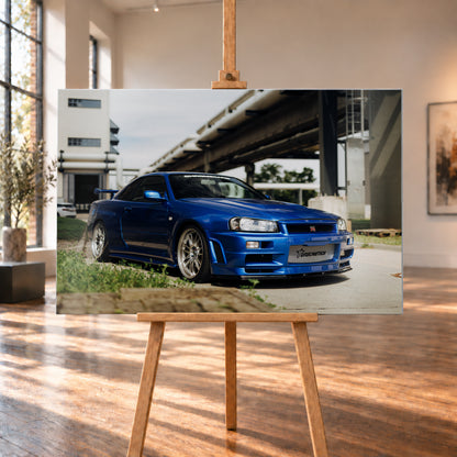 Blauer Nissan Skyline R34 GT-R in tiefer Front-Dreiviertelansicht auf Asphalt unter einer erhöhten Betonstruktur; sichtbar sind silberne Mehrspeichenräder, Heckflügel, GT-R-Emblem und Gras am Bildrand; Visualisierung als Alu gebürstet-Wandbild, auf einer Holzstaffelei in einem hellen Galerieraum.