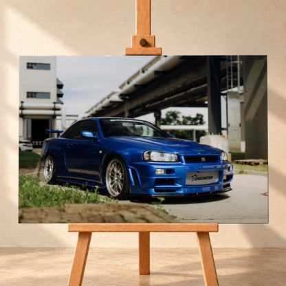 Blauer Nissan Skyline R34 GT-R in tiefer Front-Dreiviertelansicht auf Asphalt unter einer erhöhten Betonstruktur; sichtbar sind silberne Mehrspeichenräder, Heckflügel, GT-R-Emblem und Gras am Bildrand; Visualisierung als Leinwand-Wandbild, frontal auf einer Holzstaffelei.