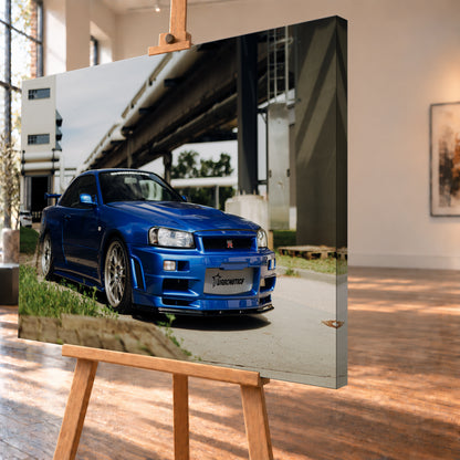 Blauer Nissan Skyline R34 GT-R in tiefer Front-Dreiviertelansicht auf Asphalt unter einer erhöhten Betonstruktur; sichtbar sind silberne Mehrspeichenräder, Heckflügel, GT-R-Emblem und Gras am Bildrand; Visualisierung als Leinwand-Wandbild, schräg von der Seite auf einer Holzstaffelei.