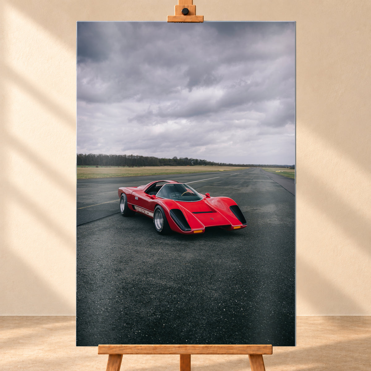 Roter Coyote X in Front-Dreiviertelansicht auf einer breiten dunklen Asphaltbahn. Das hochformatige Bild zeigt flache Wiesen, einen niedrigen Baumhorizont und einen ausgedehnten grauen Wolkenhimmel; Personen sind nicht sichtbar; Visualisierung als Alu gebürstet-Wandbild, frontal auf einer Holzstaffelei.