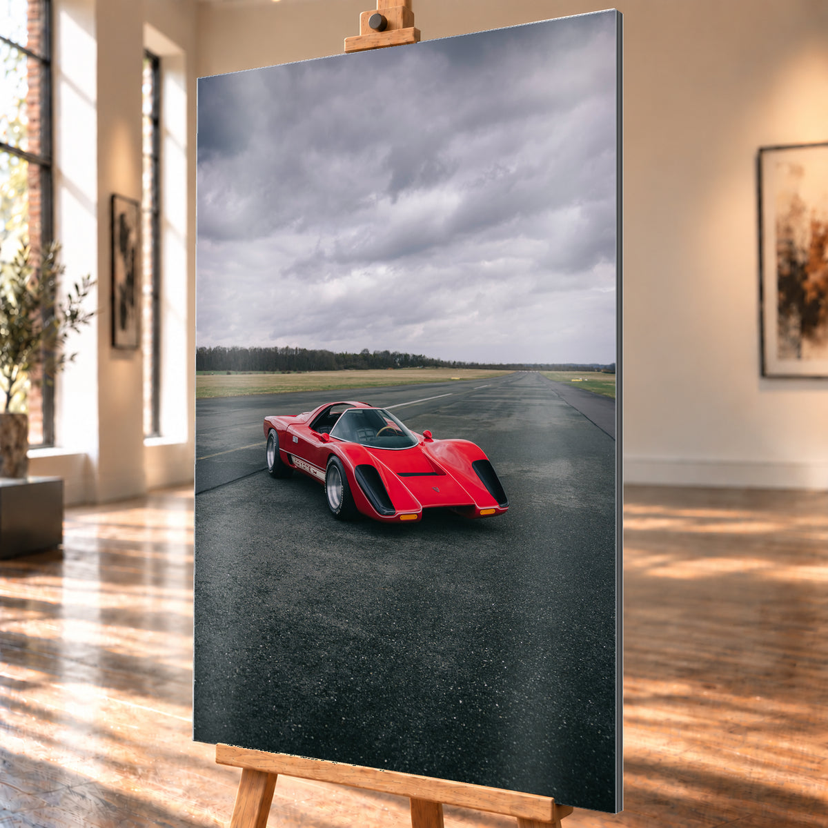 Roter Coyote X in Front-Dreiviertelansicht auf einer breiten dunklen Asphaltbahn. Das hochformatige Bild zeigt flache Wiesen, einen niedrigen Baumhorizont und einen ausgedehnten grauen Wolkenhimmel; Personen sind nicht sichtbar; Visualisierung als Alu gebürstet-Wandbild, schräg von der Seite auf einer Holzstaffelei.