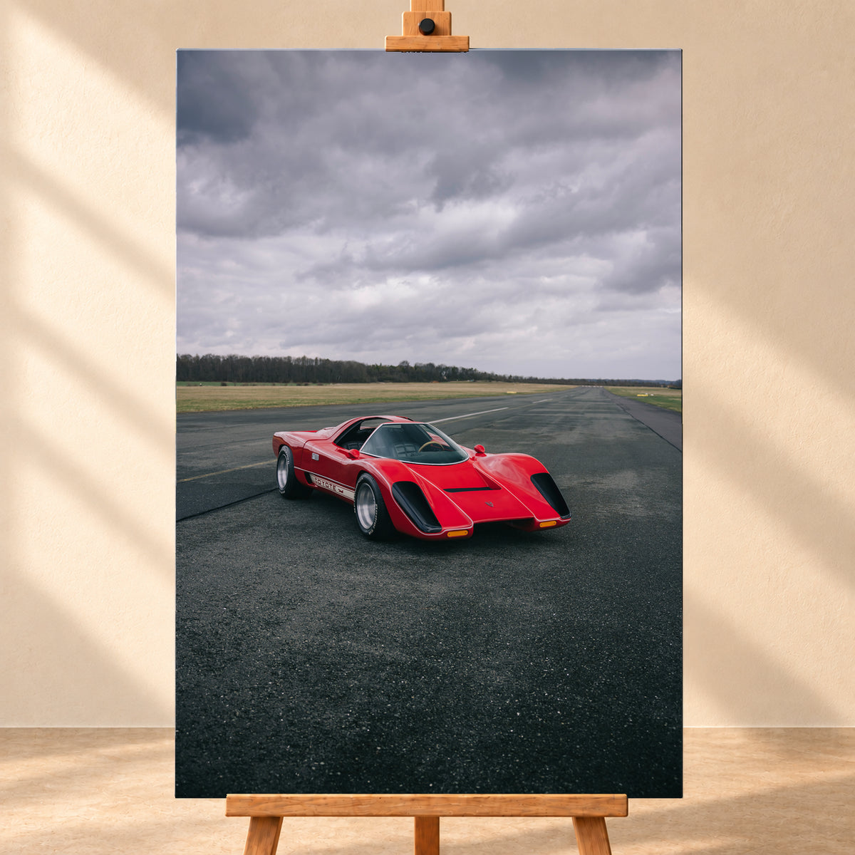 Roter Coyote X in Front-Dreiviertelansicht auf einer breiten dunklen Asphaltbahn. Das hochformatige Bild zeigt flache Wiesen, einen niedrigen Baumhorizont und einen ausgedehnten grauen Wolkenhimmel; Personen sind nicht sichtbar; Visualisierung als Leinwand-Wandbild, frontal auf einer Holzstaffelei.