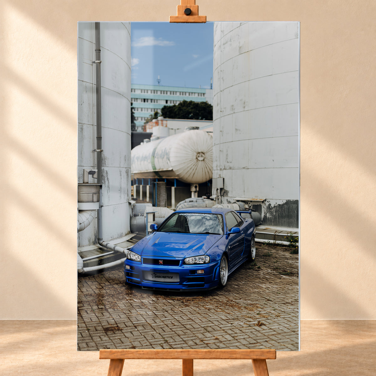 Blauer Nissan Skyline R34 GT-R in frontaler Dreiviertelansicht auf gepflastertem Industriegelände zwischen hohen weißen Behältern, Tanks und Rohrleitungen bei Tageslicht; Visualisierung als Acrylglas-Wandbild, frontal auf einer Holzstaffelei.