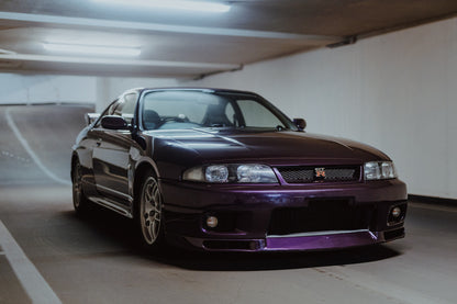 Der Nissan Skyline R33 steht tief in einer Dreiviertel-Frontansicht zwischen weißen Fahrbahnmarkierungen und kühlen Betonflächen. Originalmotiv „Midnight Moment 2“ im Querformat.