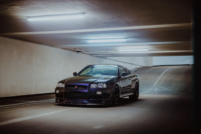 Der Nissan Skyline R34 nähert sich optisch aus einer breiten, geschwungenen Parkhausspur; das violette Auto steht im Zentrum des Lichtkegels. Originalmotiv „Midnight Focus 2“ im Querformat.