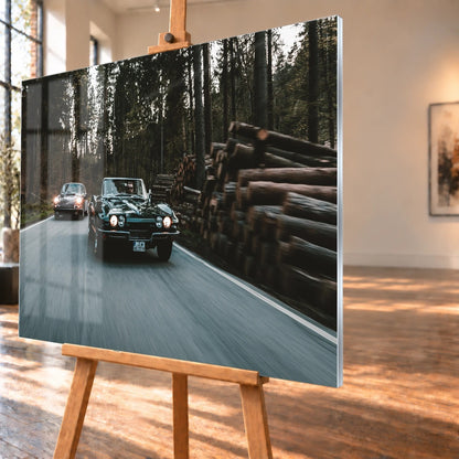 Eine grüne Corvette C2 und ein Porsche 911 S Softwindow fahren hintereinander durch einen Wald; Bewegungsunschärfe vermittelt Tempo. Motiv „Golden Light“ als Acrylglas-Wandbild, schräg von der Seite auf einer Holzstaffelei.