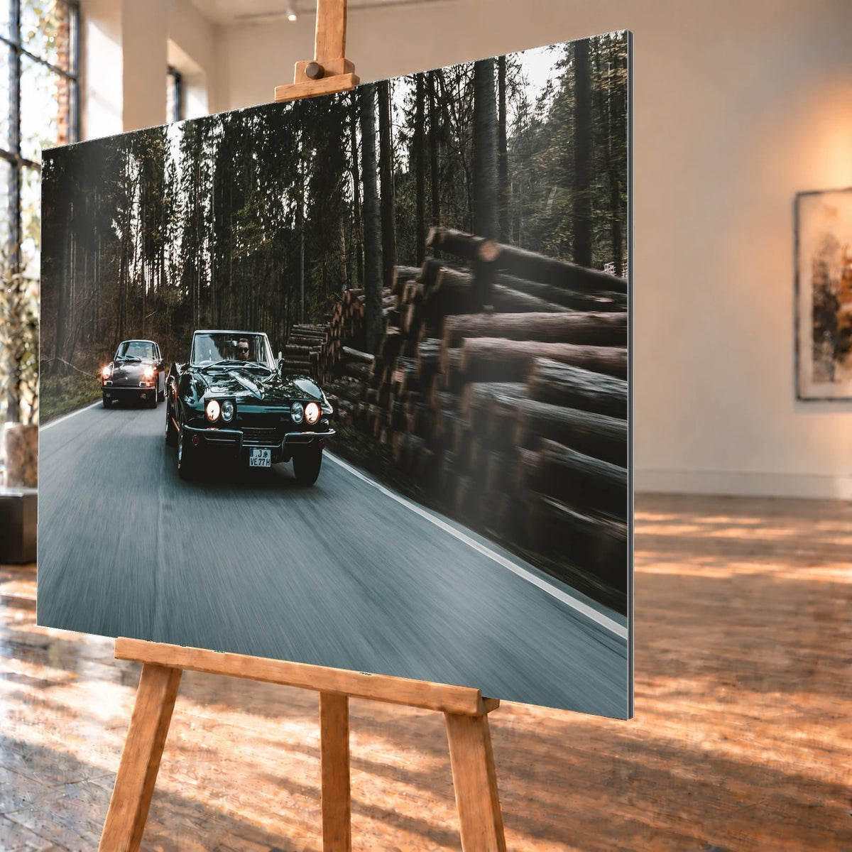 Eine grüne Corvette C2 und ein Porsche 911 S Softwindow fahren hintereinander durch einen Wald; Bewegungsunschärfe vermittelt Tempo. Motiv „Golden Light“ als Alu gebürstet-Wandbild, schräg von der Seite auf einer Holzstaffelei.