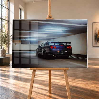 Der Nissan Skyline R34 steht schräg von hinten unter langen Deckenleuchten; Spoiler und rote Rundlichter führen die Perspektive. Motiv „Skyline Presence 2“ als Acrylglas-Wandbild, auf einer Holzstaffelei in einem hellen Galerieraum.