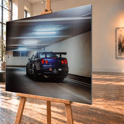 Der Nissan Skyline R34 steht schräg von hinten unter langen Deckenleuchten; Spoiler und rote Rundlichter führen die Perspektive. Motiv „Skyline Presence 2“ als Alu gebürstet-Wandbild, schräg von der Seite auf einer Holzstaffelei.