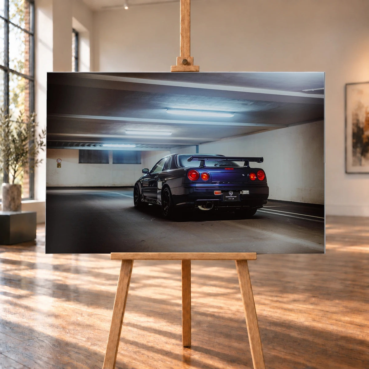 Der Nissan Skyline R34 steht schräg von hinten unter langen Deckenleuchten; Spoiler und rote Rundlichter führen die Perspektive. Motiv „Skyline Presence 2“ als Alu gebürstet-Wandbild, auf einer Holzstaffelei in einem hellen Galerieraum.