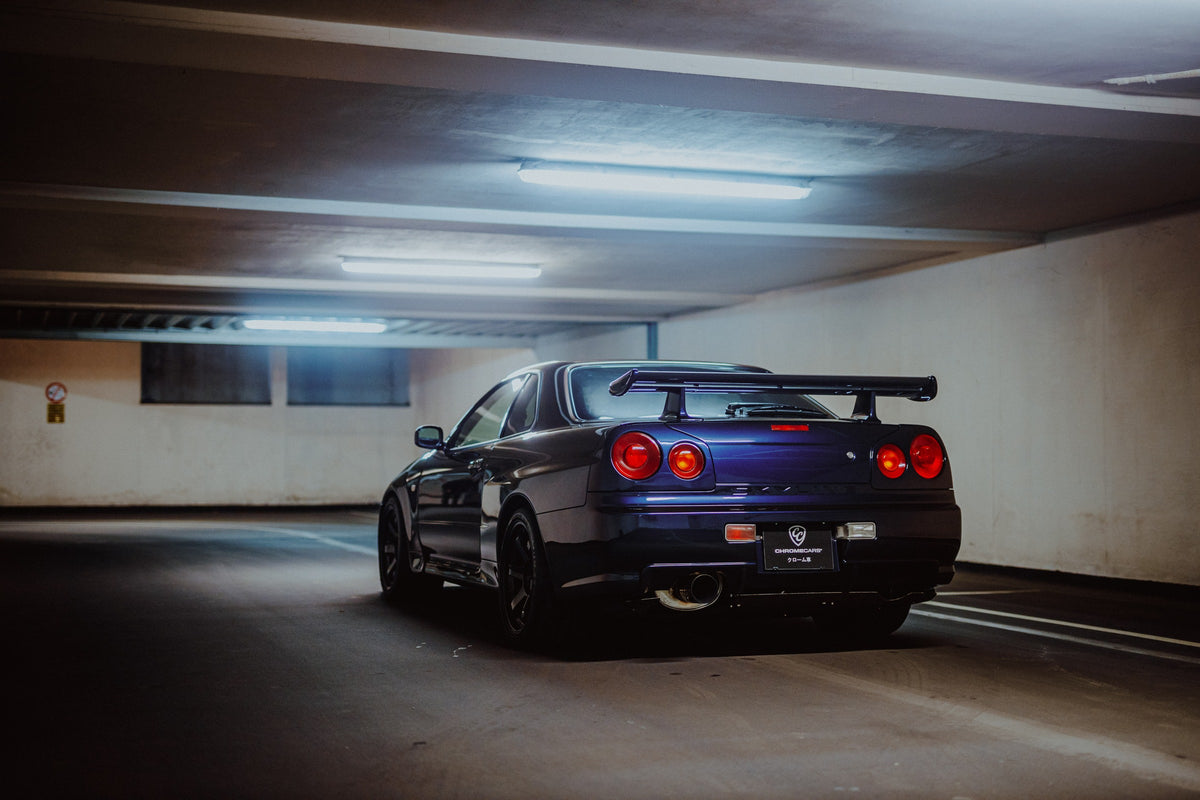 Der Nissan Skyline R34 steht schräg von hinten unter langen Deckenleuchten; Spoiler und rote Rundlichter führen die Perspektive. Originalmotiv „Skyline Presence 2“ im Querformat.