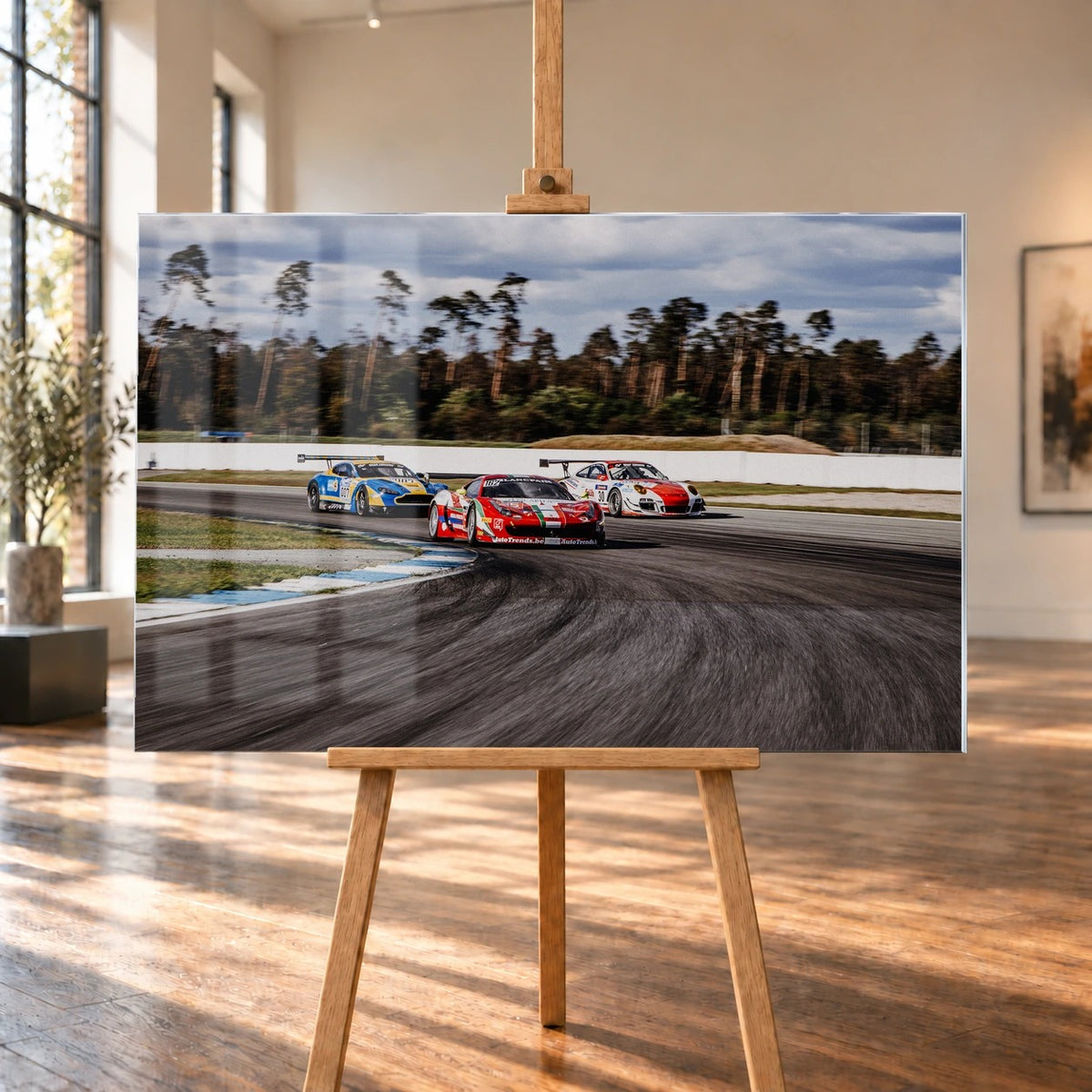 Aston Martin, Porsche und Ferrari GT3 fahren dicht gestaffelt durch eine Kurve; die Mehrwagen-Szene zeigt echtes Renngeschehen. Motiv „Racing Echo“ als Acrylglas-Wandbild, auf einer Holzstaffelei in einem hellen Galerieraum.