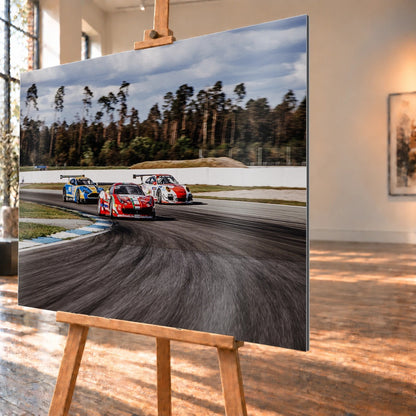 Aston Martin, Porsche und Ferrari GT3 fahren dicht gestaffelt durch eine Kurve; die Mehrwagen-Szene zeigt echtes Renngeschehen. Motiv „Racing Echo“ als Alu gebürstet-Wandbild, schräg von der Seite auf einer Holzstaffelei.