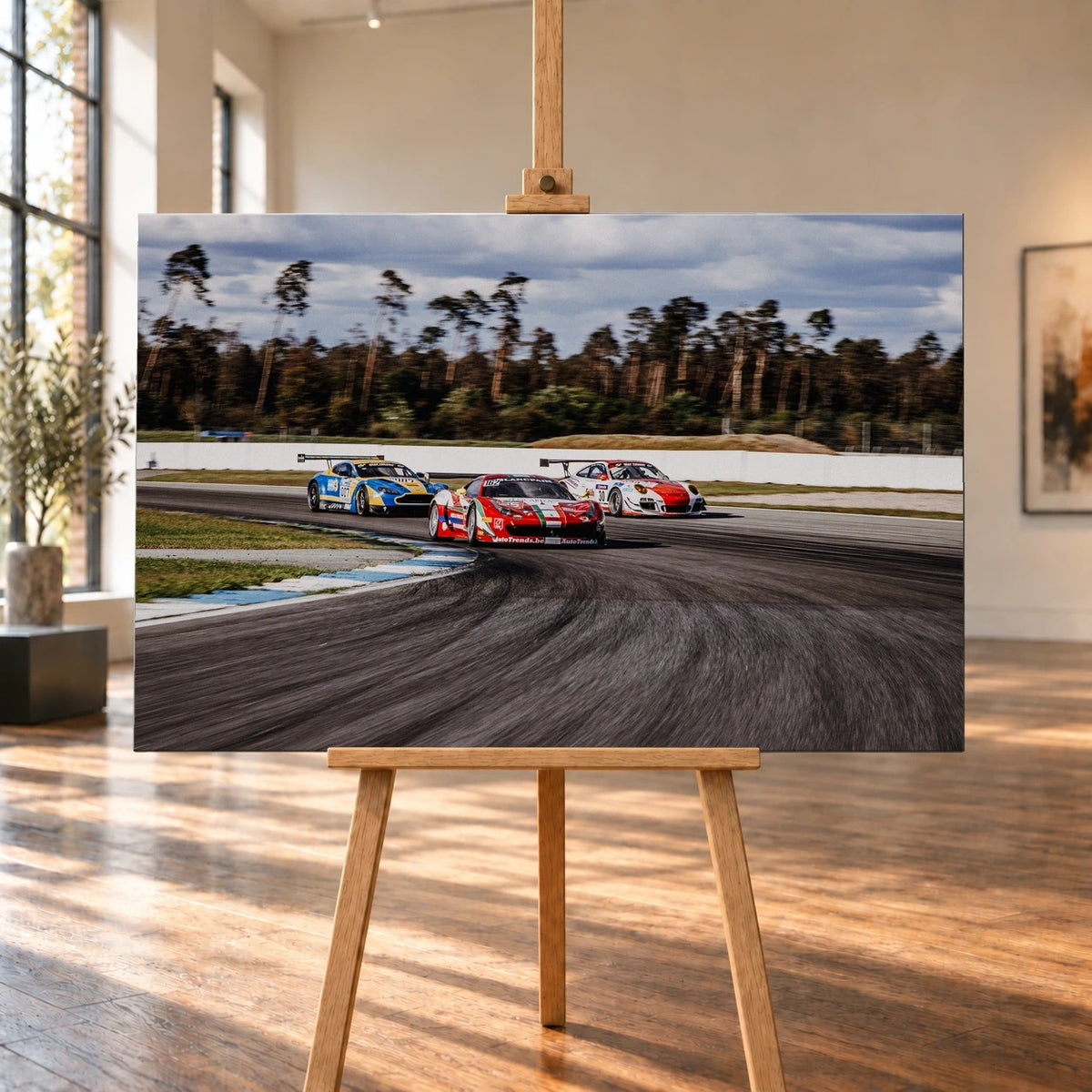 Aston Martin, Porsche und Ferrari GT3 fahren dicht gestaffelt durch eine Kurve; die Mehrwagen-Szene zeigt echtes Renngeschehen. Motiv „Racing Echo“ als Leinwand-Wandbild, auf einer Holzstaffelei in einem hellen Galerieraum.