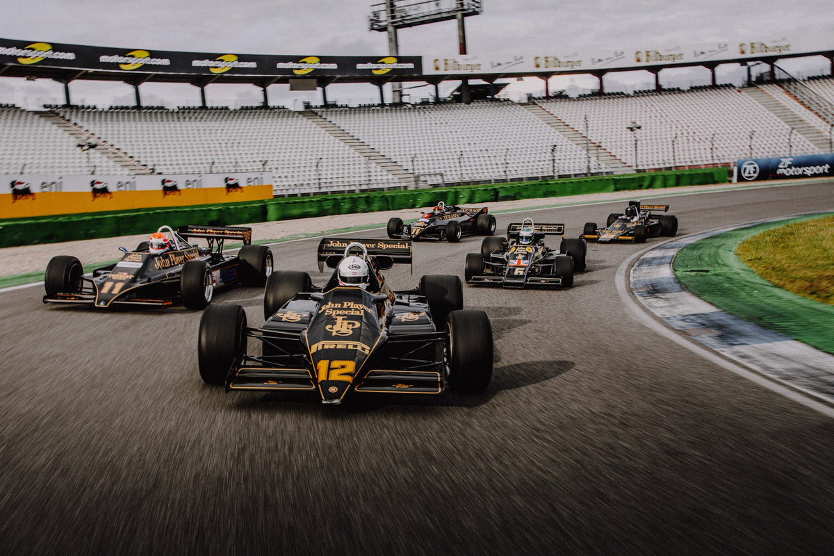 Fünf historische Lotus-Formelwagen fahren als Gruppe durch eine Rennstreckenkurve; schwarze Gold-Lackierungen und Startnummern sind deutlich erkennbar. Originalmotiv „Trackside Story“ im Querformat.