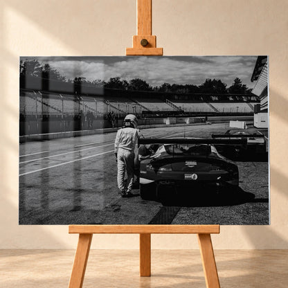 Der Aston Martin Vantage V12 GT3 Billy steht von hinten in der Boxengasse; eine Person lehnt am geöffneten Heckbereich und gibt dem Bild Maßstab. Motiv „Racing Frame“ als Acrylglas-Wandbild, frontal auf einer Holzstaffelei.