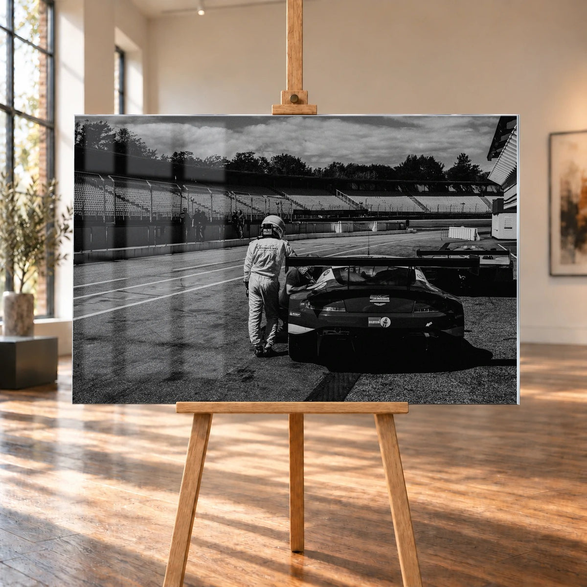 Der Aston Martin Vantage V12 GT3 Billy steht von hinten in der Boxengasse; eine Person lehnt am geöffneten Heckbereich und gibt dem Bild Maßstab. Motiv „Racing Frame“ als Acrylglas-Wandbild, auf einer Holzstaffelei in einem hellen Galerieraum.