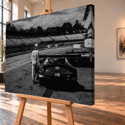 Der Aston Martin Vantage V12 GT3 Billy steht von hinten in der Boxengasse; eine Person lehnt am geöffneten Heckbereich und gibt dem Bild Maßstab. Motiv „Racing Frame“ als Leinwand-Wandbild, schräg von der Seite auf einer Holzstaffelei.