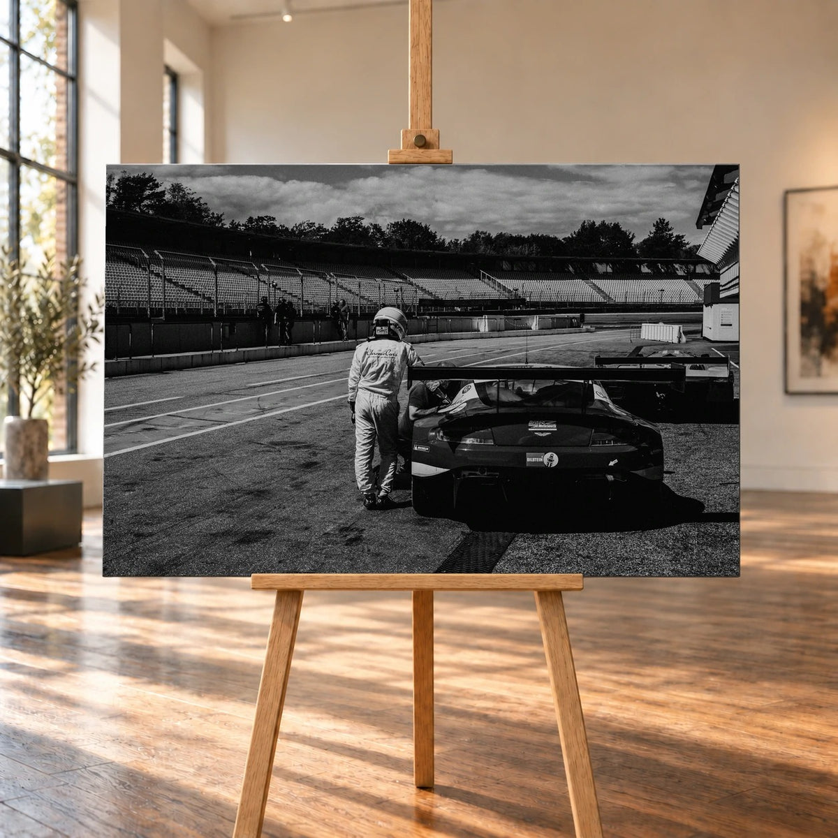 Der Aston Martin Vantage V12 GT3 Billy steht von hinten in der Boxengasse; eine Person lehnt am geöffneten Heckbereich und gibt dem Bild Maßstab. Motiv „Racing Frame“ als Leinwand-Wandbild, auf einer Holzstaffelei in einem hellen Galerieraum.