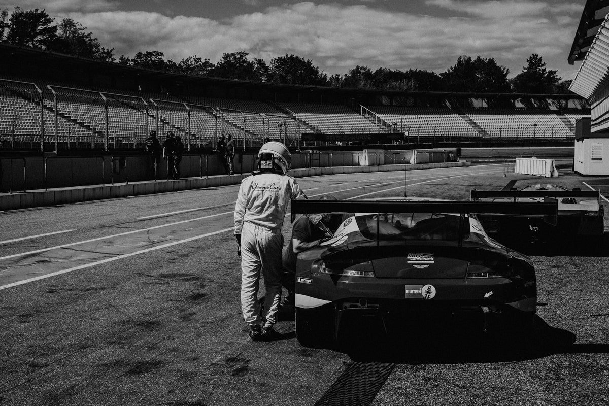 Der Aston Martin Vantage V12 GT3 Billy steht von hinten in der Boxengasse; eine Person lehnt am geöffneten Heckbereich und gibt dem Bild Maßstab. Originalmotiv „Racing Frame“ im Querformat.