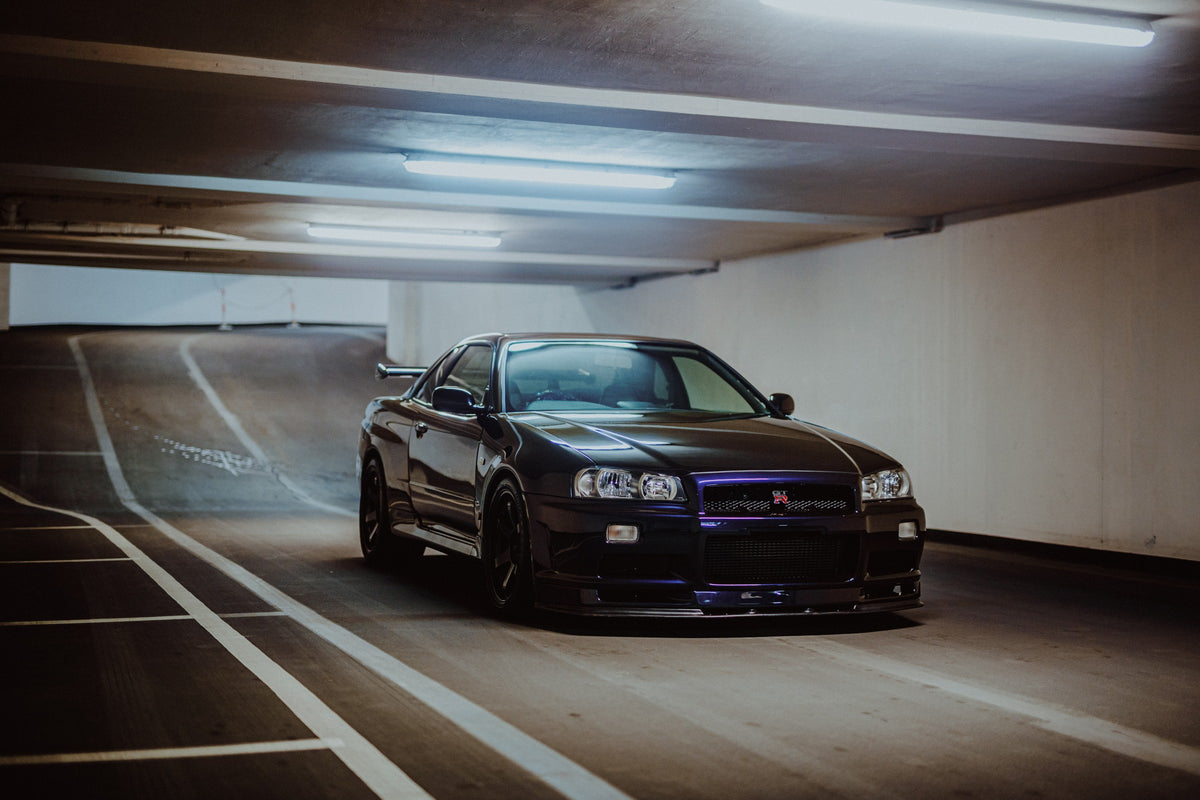 Der Nissan Skyline R34 steht tief und breit in der Kurve eines Parkhauses; Front, Seitenschweller und violette Akzente sind klar sichtbar. Originalmotiv „Violet Story“ im Querformat.